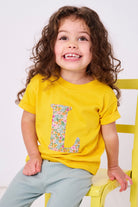 a smiling 2 year old girl wearing a personalised kids yellow t-shirt with an initial cut from poppy forest liberty floral print on the front