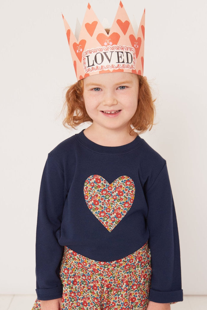 A 4 year old girl wearing a children's valentines outfit. A navy cotton t-shirt with a Liberty floral heart sewn on the front and a crown that says 'loved' on it.