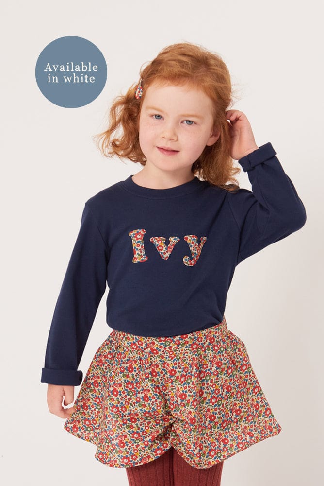 A 4 year old girl wearing a personalised navy cotton t-shirt with her name on the front. Her name is appliquéd in lowercase letters in Liberty floral fabric.