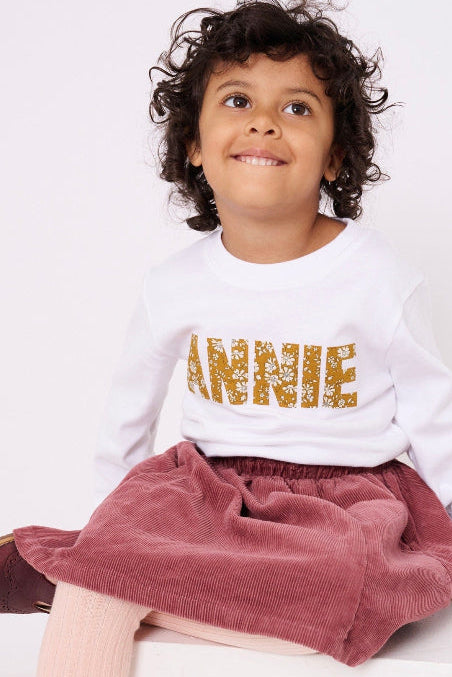 A young girl wearing a white personalised t-shirt with her name on the front cut from Capel Mustard liberty print.
