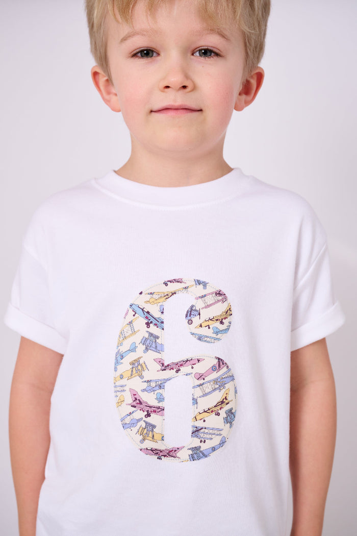 A 6 year old boy wearing a white cotton t-shirt with a number 6 sewn on the front made from Tom's Jet plane Liberty fabric.