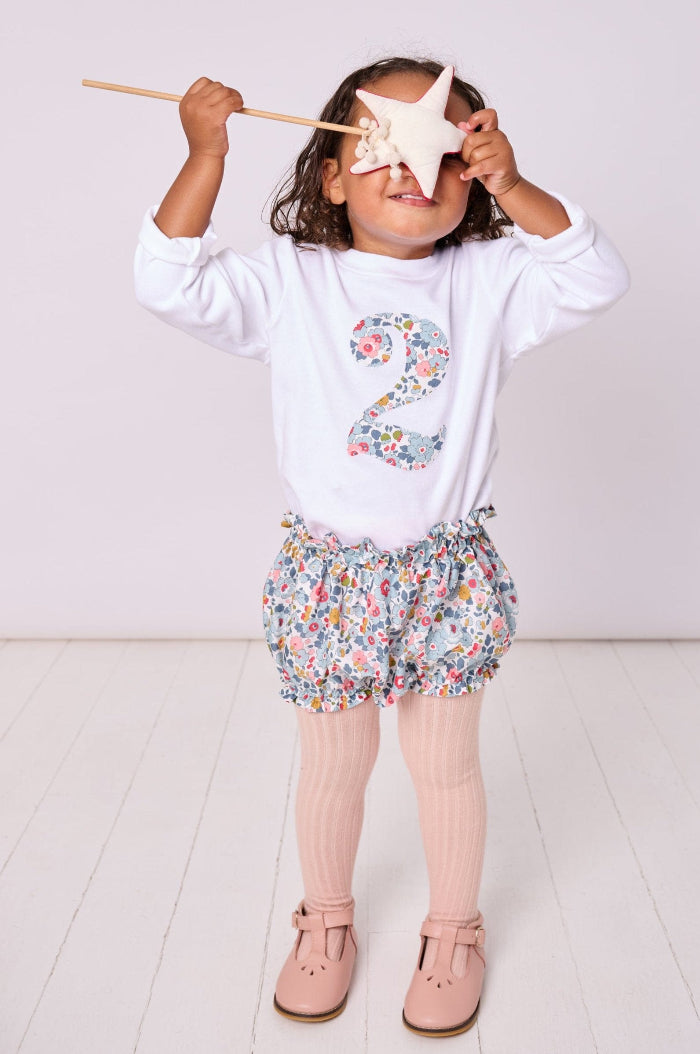 A child wearing a white t-shirt with the number 2 in Betsy Grey Liberty Print, paired with floral bloomers, holding a wand and standing against a white background.