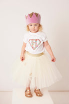 A 3 year old girl wearing a personalised  white t-shirt with a superhero motif initial made from floral Liberty print fabric.