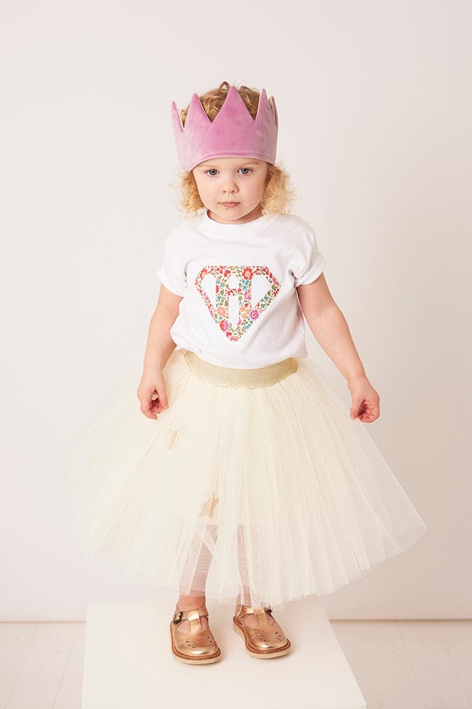A 3 year old girl wearing a personalised  white t-shirt with a superhero motif initial made from floral Liberty print fabric.
