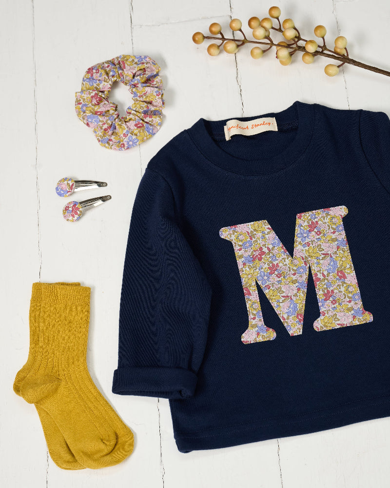 A navy blue long-sleeve t-shirt with a large Liberty floral monogram letter 'M' on the front, styled in a flat lay with a matching floral scrunchie, two matching hair clips, a pair of mustard yellow socks, and a decorative berry branch on a white wood plank background.