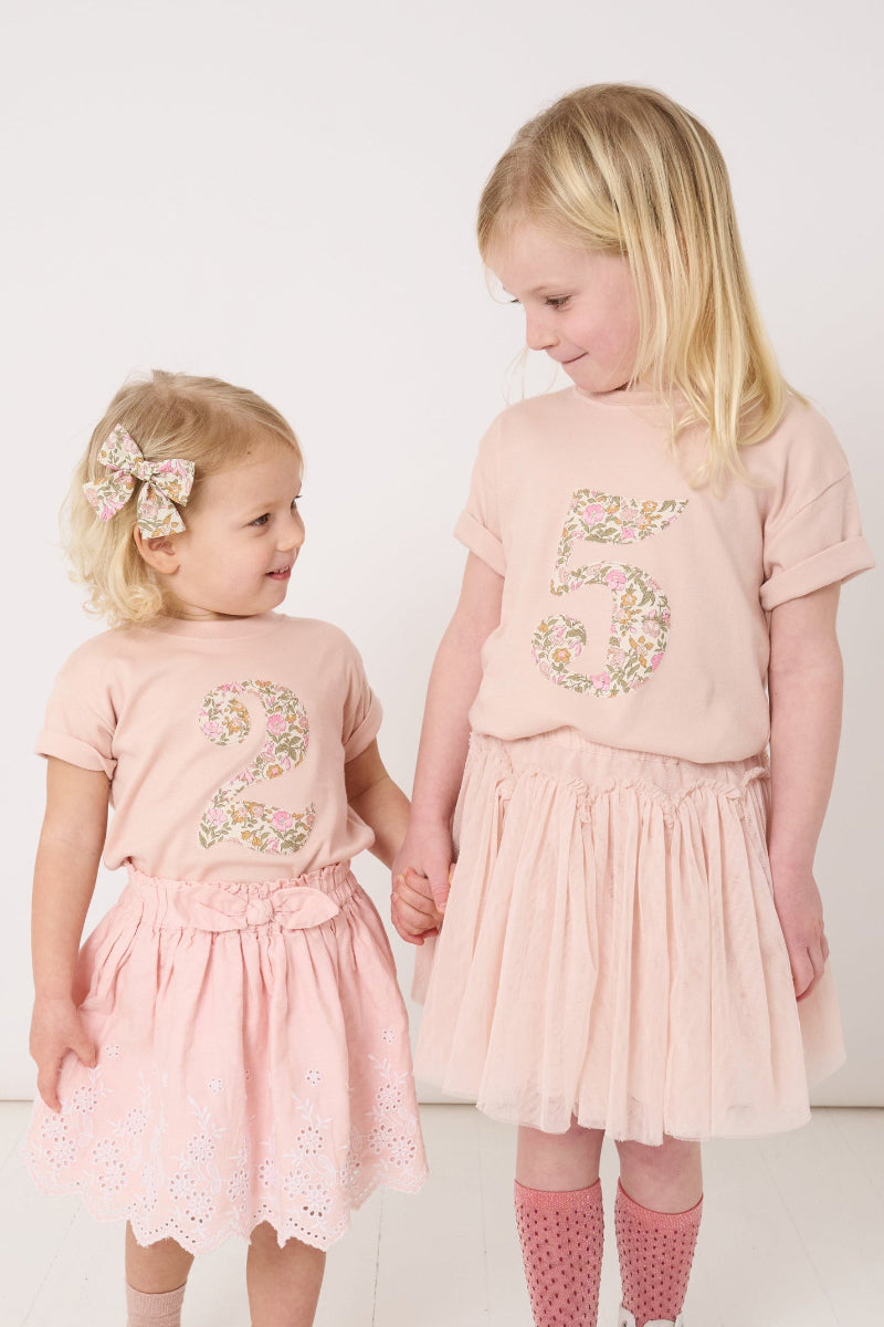 Two young girls in matching pink floral birthday t-shirts, one with a number 2 and one with a number 5 standing side by side on a white background.