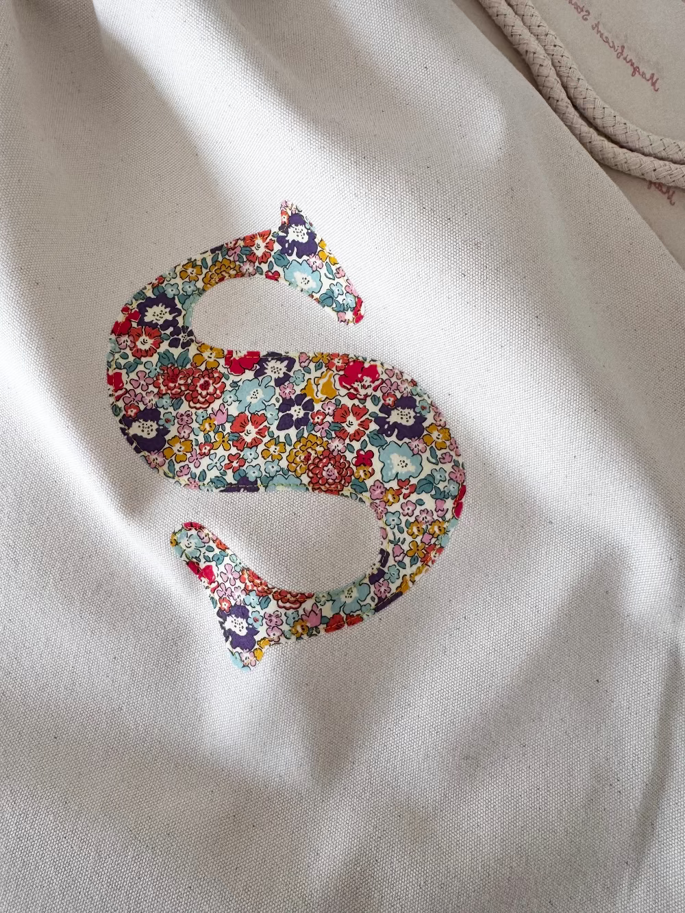 A cream canvas drawstring backpack with a personalised Liberty floral initial sewn on the front