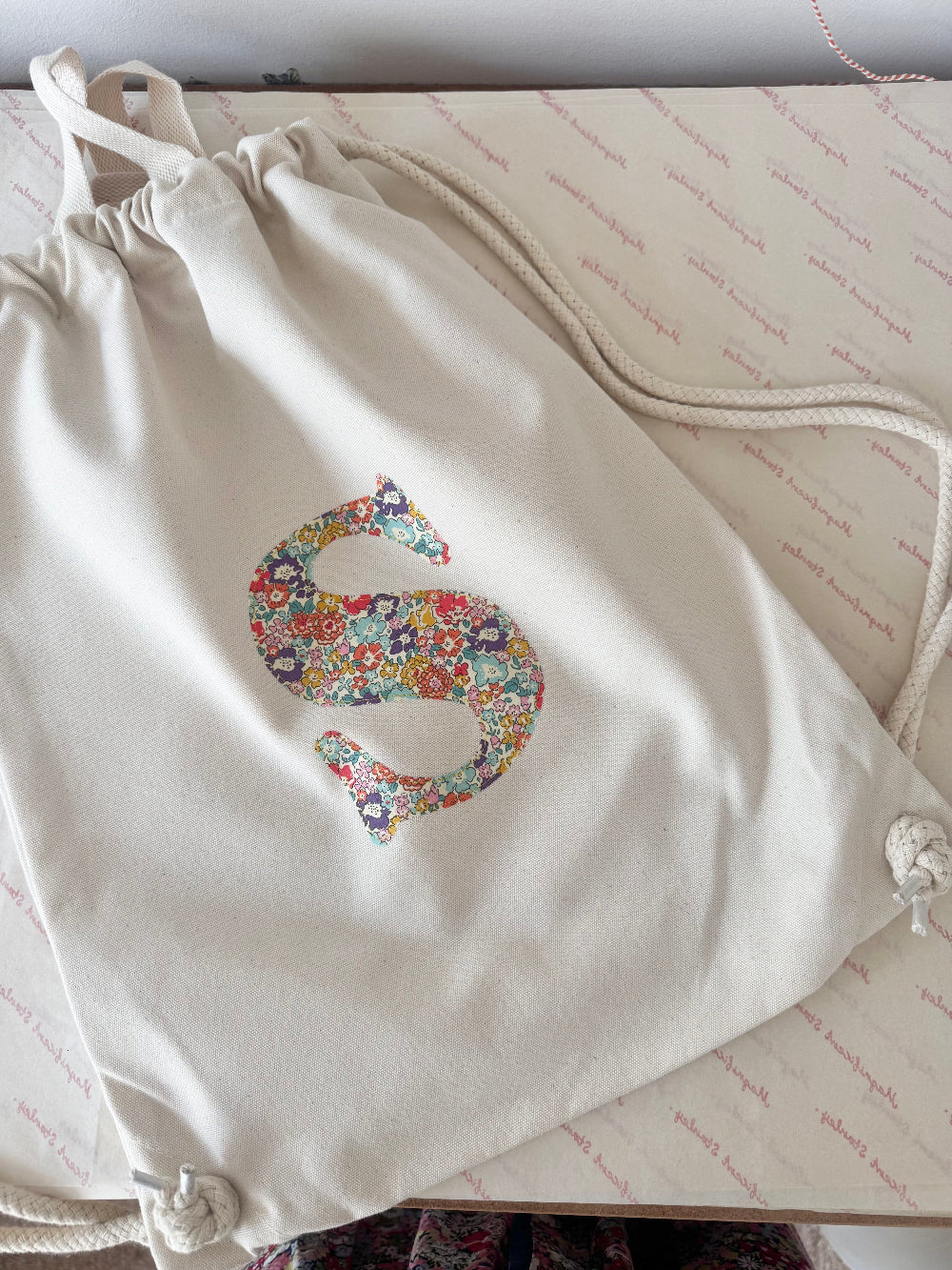 A cream canvas drawstring backpack with a personalised Liberty floral initial sewn on the front