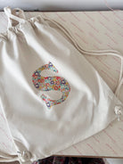 A cream canvas drawstring backpack with a personalised Liberty floral initial sewn on the front