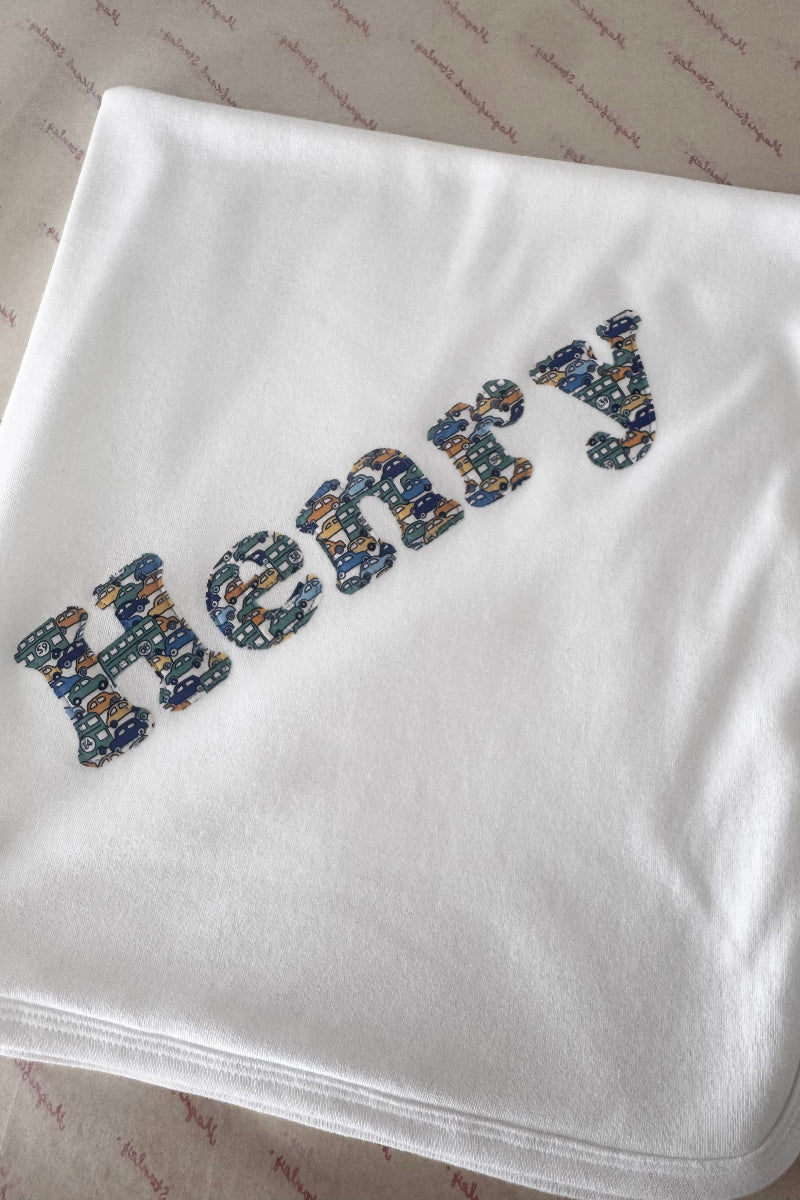 A white baby blanket or swaddle with the name 'Henry' embroidered in a blue Liberty car pattern, laid flat on a patterned beige fabric background.