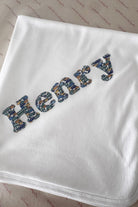 A white baby blanket or swaddle with the name 'Henry' embroidered in a blue Liberty car pattern, laid flat on a patterned beige fabric background.