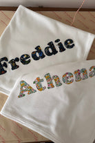 Two white baby blankets or swaddle blankets laid flat on a patterned surface. The top blanket is embroidered with the name 'Freddie' in dark Liberty space patterned letters, and the bottom blanket is embroidered with the name 'Athena' in multicoloured floral Liberty letters.