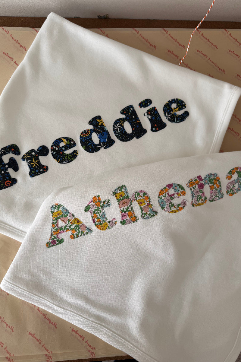 Two white baby blankets or swaddle blankets laid flat on a patterned surface. The top blanket is embroidered with the name 'Freddie' in dark Liberty space patterned letters, and the bottom blanket is embroidered with the name 'Athena' in multicoloured floral Liberty letters.