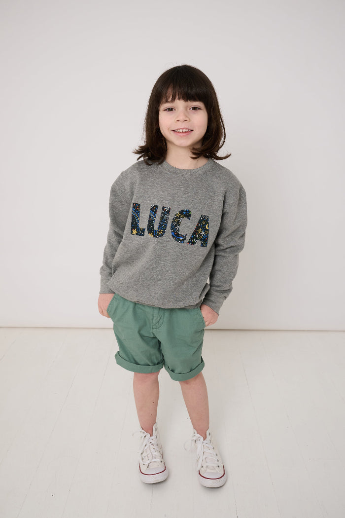 Child wearing a grey sweatshirt with 'LUCA' in colourful letters in space liberty print.
