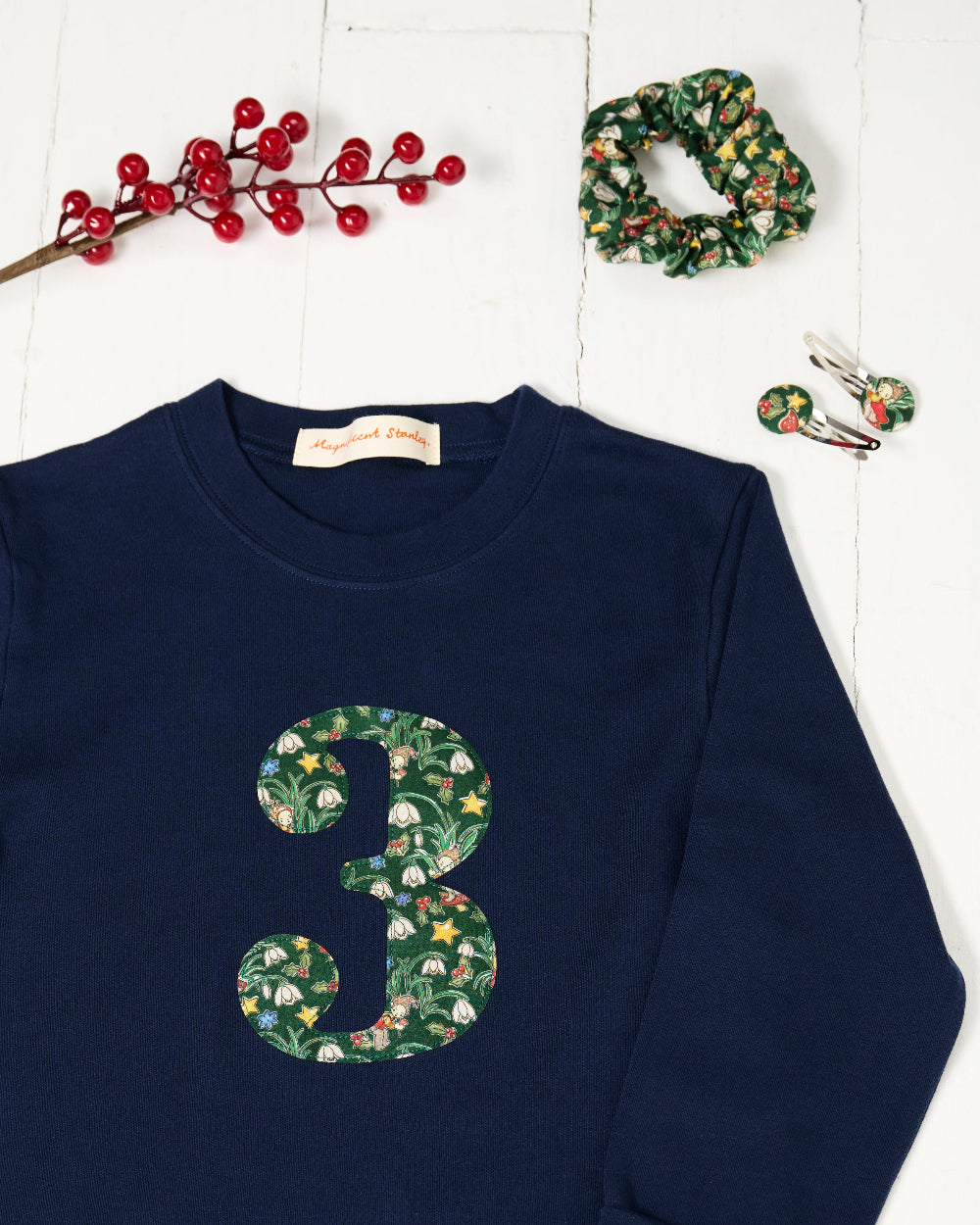 A navy blue kids cotton t-shirt laid flat on a white wood background, featuring a large number '3' on the front filled with a green holiday-themed pattern of pinecones, ornaments, and snowdrops called Liberty Elves. Styled flat-lay props include red berries, a green patterned scrunchie, and matching hair clips.