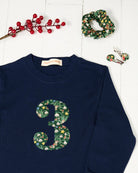 A navy blue kids cotton t-shirt laid flat on a white wood background, featuring a large number '3' on the front filled with a green holiday-themed pattern of pinecones, ornaments, and snowdrops called Liberty Elves. Styled flat-lay props include red berries, a green patterned scrunchie, and matching hair clips.