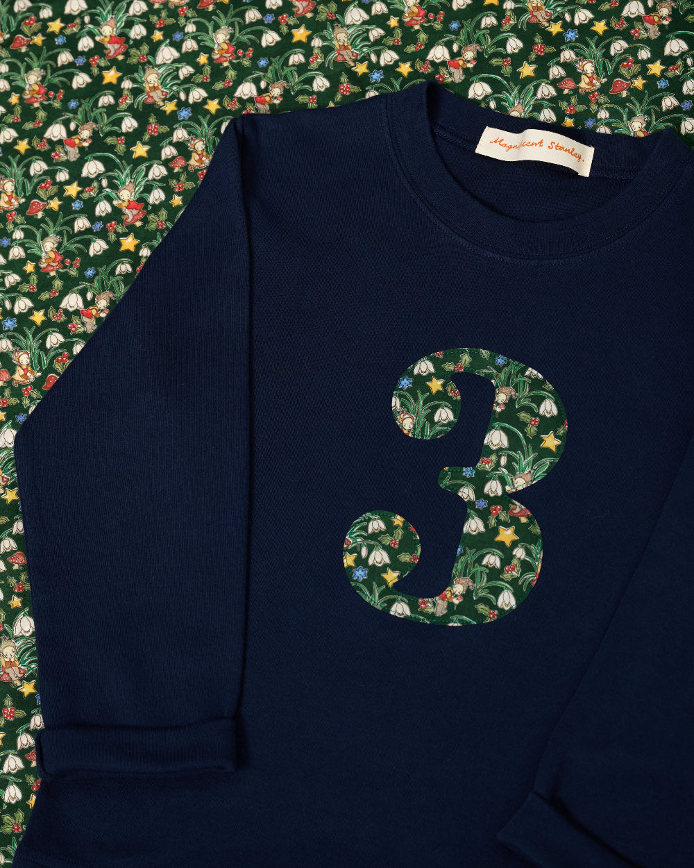 A navy blue kids cotton t-shirt laid flat on a a Liberty fabric background, featuring a large number '3' on the front filled with a green holiday-themed pattern of pinecones, ornaments, and snowdrops called Liberty Eleves. 