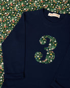 A navy blue kids cotton t-shirt laid flat on a a Liberty fabric background, featuring a large number '3' on the front filled with a green holiday-themed pattern of pinecones, ornaments, and snowdrops called Liberty Eleves. 