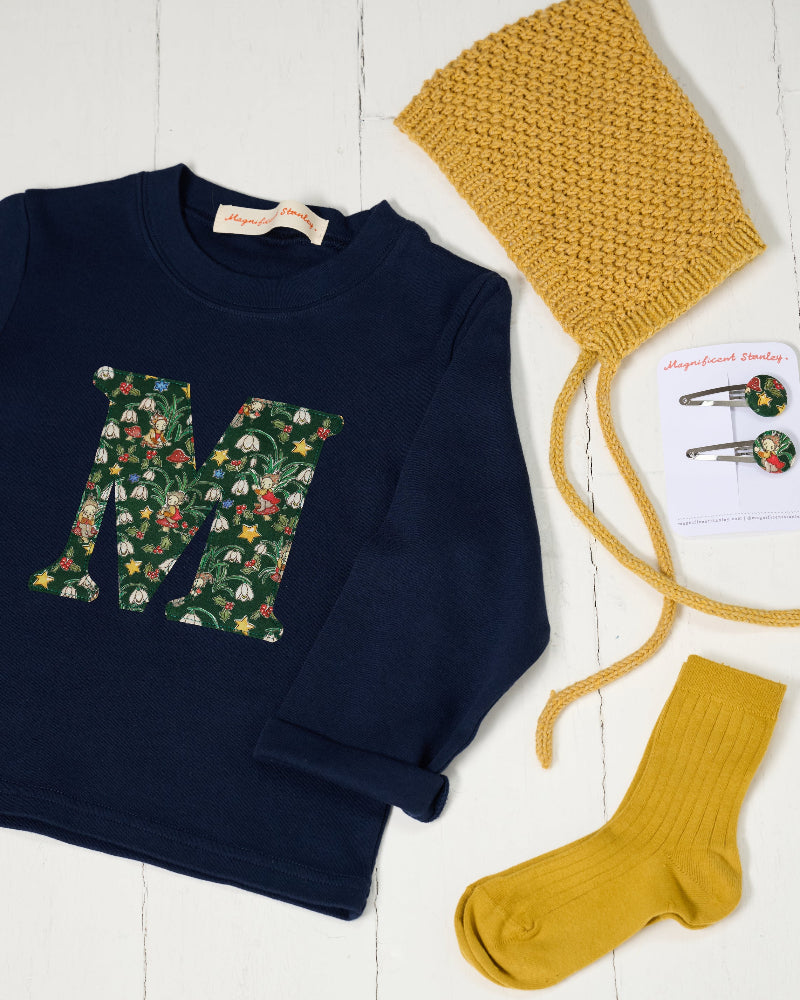 A styled flat-lay of a children's outfit set on a white wood background, featuring a navy blue long-sleeve t-shirt with a large floral monogram 'M' on the front in Liberty Elves print featuring holly, snowdrops, stars and elves. It also has a mustard yellow knit bonnet with ties, a pair of matching mustard yellow socks, and a pair of Liberty floral hair clips on a card.