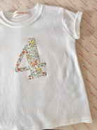 A white short-sleeve baby tee with a large floral-patterned number 4 on the front, laid flat on a patterned fabric background. The inner neck label reads 'Palmie Daisy'.