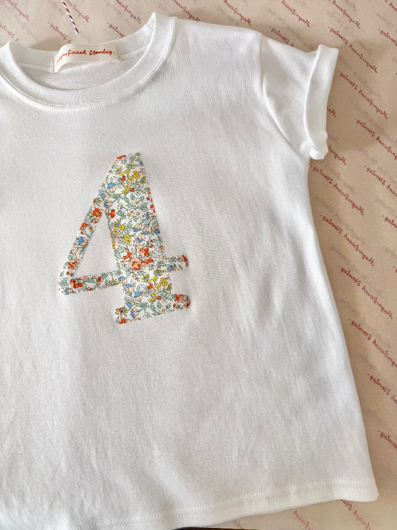 A white short-sleeve baby tee with a large floral-patterned number 4 on the front, laid flat on a patterned fabric background. The inner neck label reads 'Palmie Daisy'.