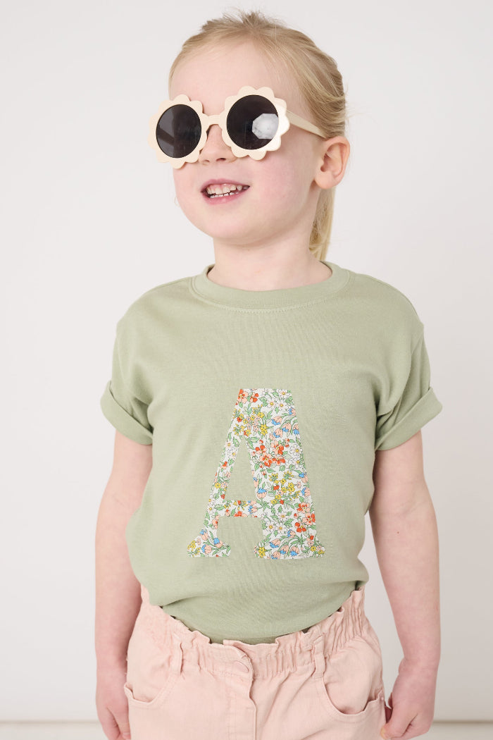 a 5 year old girl wearing a personalised kids sage green t-shirt with his initial cut from Nazanin liberty print on the front.