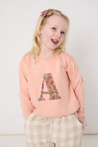 Young girl wearing a pink sweatshirt with a Liberty floral letter 'A' design and plaid trousers on a white background