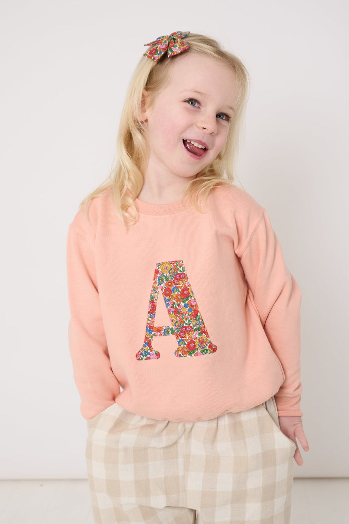 Young girl wearing a pink sweatshirt with a Liberty floral letter 'A' design and plaid trousers on a white background