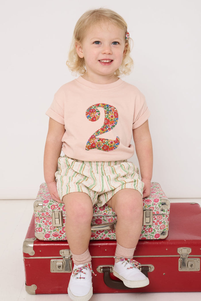 Child sitting on a red suitcase wearing a pink shirt with the number 2 and floral Liberty design, white shorts, and white shoes.