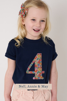 A young blonde girl wearing a navy blue t-shirt with a Liberty floral patterned number 4 on the front, paired with light pink pants. She has a matching floral hair bow.