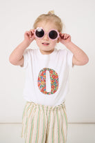 A 2 year old girl wearing a summery personalised white cotton t-shirt with her initial cut from Nell Annie and May floral liberty fabric.