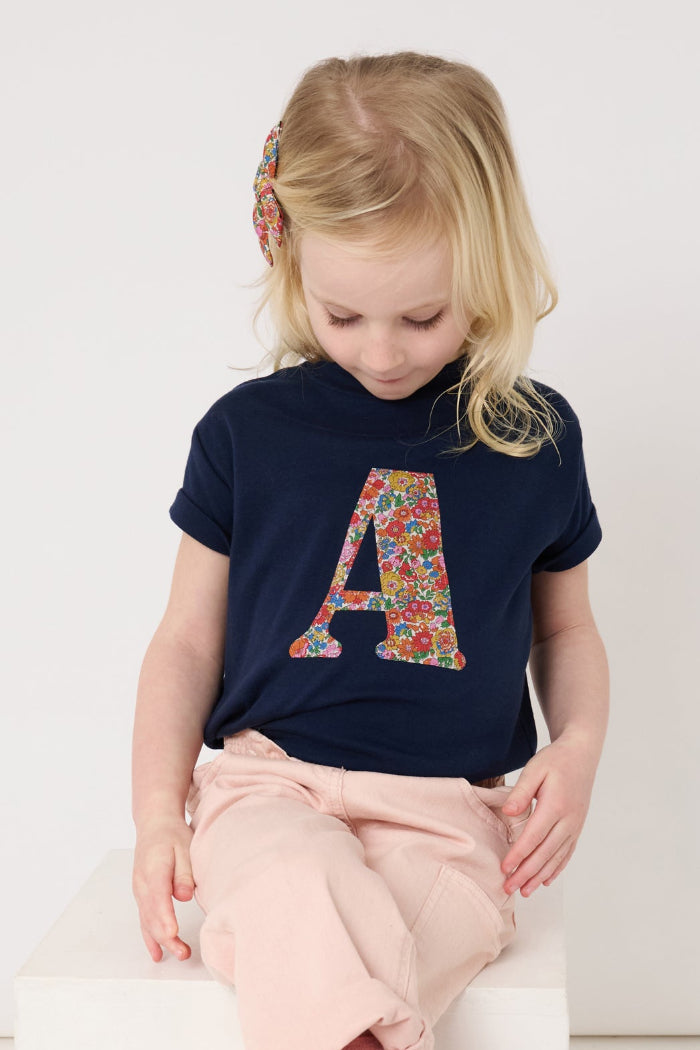 a 4 year old girl wearing a personalised kids navy t-shirt with an initial cut from nell annie and may liberty floral print on the front.
