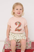 A 2 year old girl wearing a pale pink t-shirt with a Liberty Nell Annie and May floral number '2' on the front