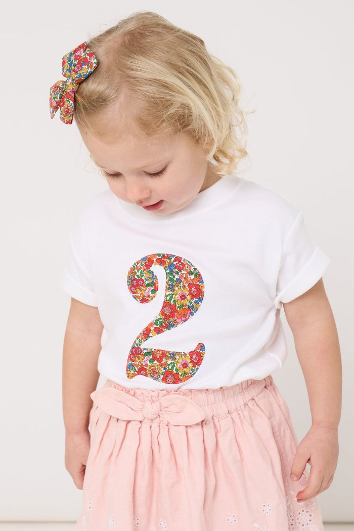 A 2 year old girl wearing a white cotton birthday t-shirt with the number 2 sewn on the front cut from liberty Nell Annie and May fabric.