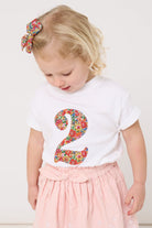 A 2 year old girl wearing a white cotton birthday t-shirt with the number 2 sewn on the front cut from liberty Nell Annie and May fabric.