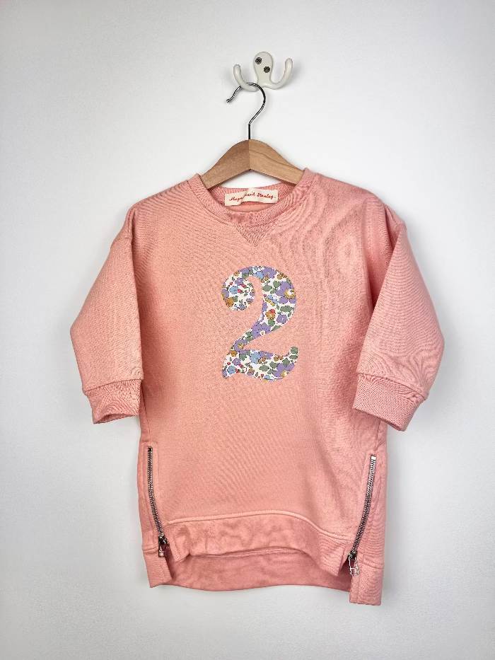 A pink crewneck sweatshirt dress hanging on a wooden hanger against a white wall. The sweatshirt features a large number '2' on the chest filled with a purple, blue, and green Liberty Betsy floral pattern, and silver zipper details on the sides.