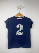 A navy blue short-sleeve t-shirt hanging on a wooden hanger from a wall hook. The shirt features a large number '2' printed on the front in a light blue and white liberty ships patterned fabric.