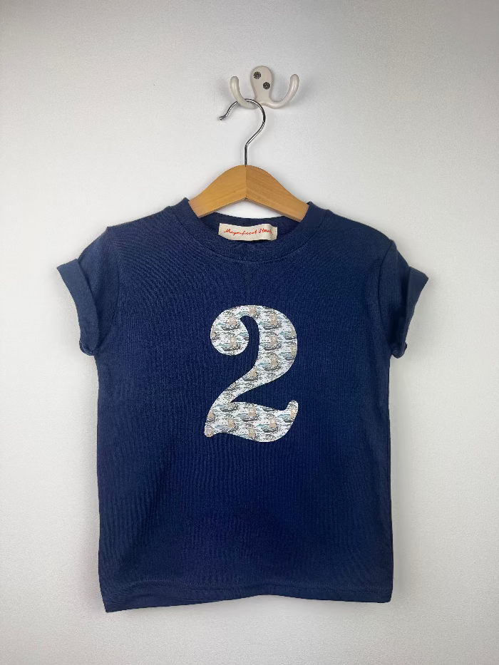 A navy blue short-sleeve t-shirt hanging on a wooden hanger from a wall hook. The shirt features a large number '2' printed on the front in a light blue and white liberty ships patterned fabric.