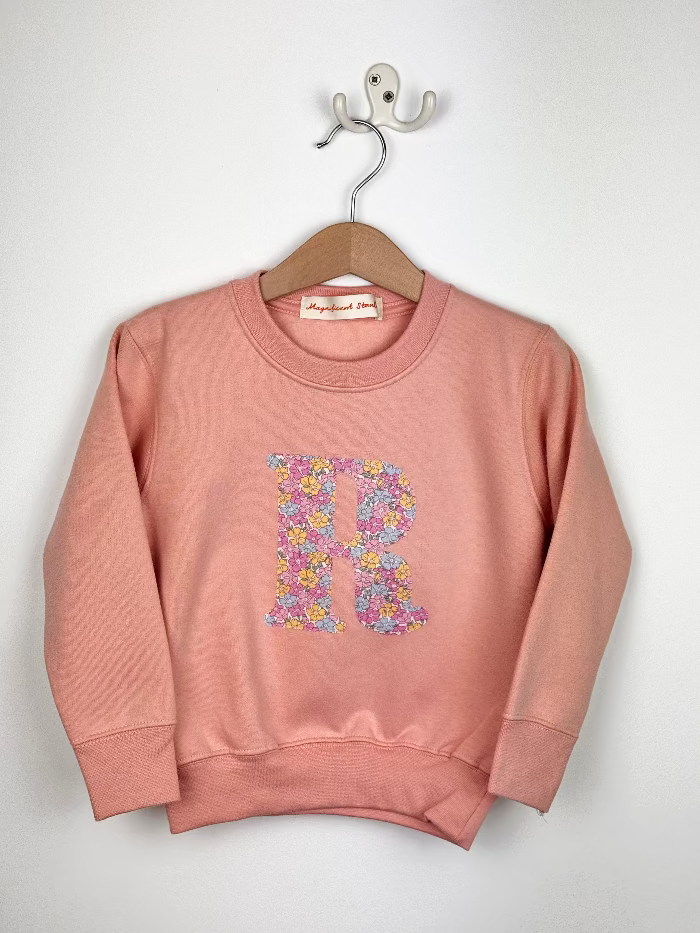 Pink sweatshirt with floral letter 'R' on a hanger against a white background