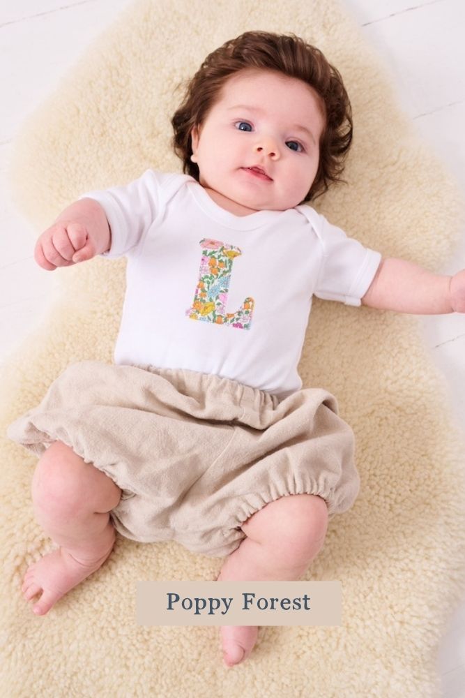 A smiling newborn baby wearing a personalised white bodysuit with an initial on the front cut from Liberty floral Poppy Forest fabric.