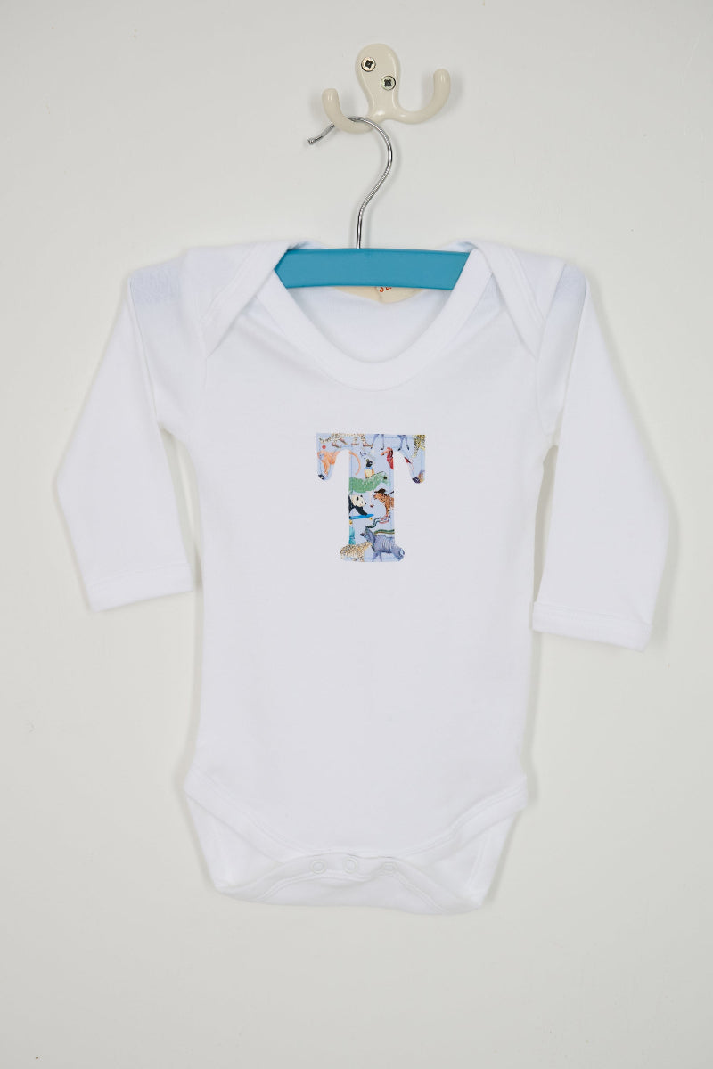 A white long-sleeve baby bodysuit hanging on a metal hanger, featuring a Quey 2 Liberty print graphic letter 'T' on the chest, set against a plain off-white wall.