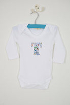 A white long-sleeve baby bodysuit hanging on a metal hanger, featuring a Quey 2 Liberty print graphic letter 'T' on the chest, set against a plain off-white wall.