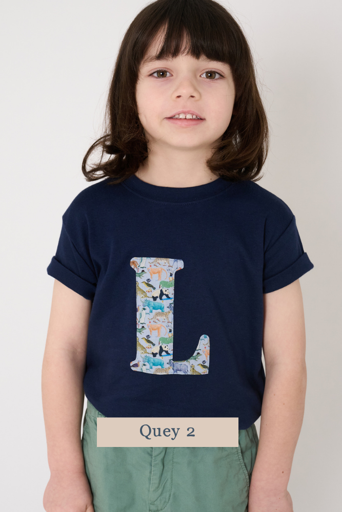 A young boy with dark brown hair is wearing a navy blue t-shirt featuring a large Liberty Queue print letter 'L' on the front. The print features zoo animals.