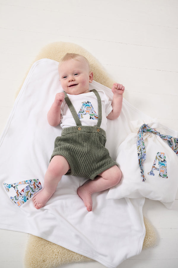 A newborn baby laying on a Personalised Cotton Baby Blanket with their initial sewn on it made from zoo animal liberty print. worn with matching babygro and gift bag.