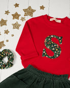 A festive children's outfit set featuring a bright red long-sleeve top with a large green floral monogram letter 'S' on the chest made from Liberty Elves print, paired with a dark green corduroy skirt and a matching green floral scrunchie, styled with gold glitter star decorations on a white background.