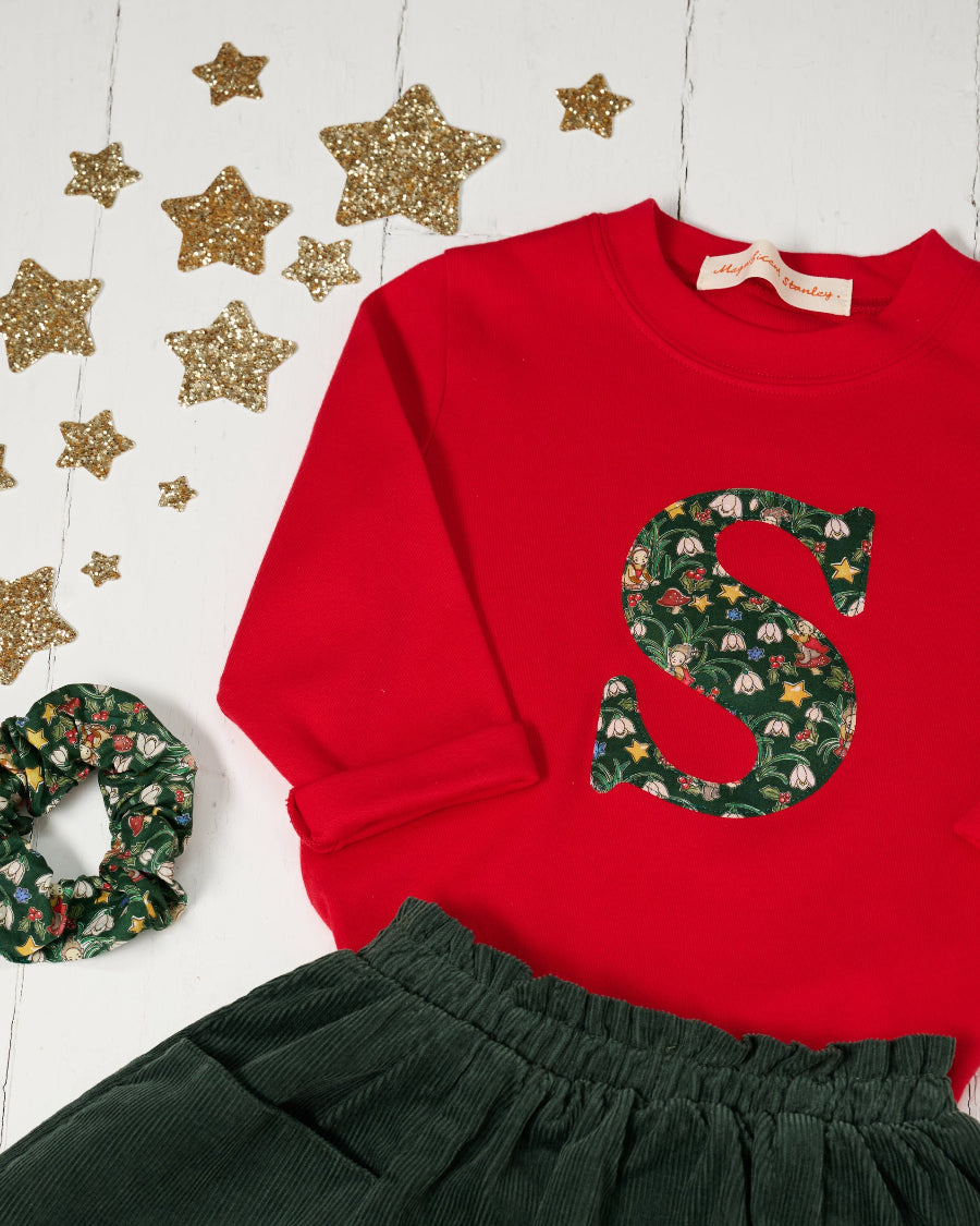 A festive children's outfit set featuring a bright red long-sleeve top with a large green floral monogram letter 'S' on the chest made from Liberty Elves print, paired with a dark green corduroy skirt and a matching green floral scrunchie, styled with gold glitter star decorations on a white background.