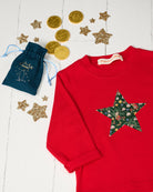A red long-sleeve sweatshirt with a large star-shaped patch on the chest featuring a Christmas Liberty Elves print, styled on a white wood background with gold coins, glitter stars, and a small blue drawstring pouch.