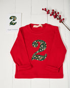 A red long-sleeve children's top with a large floral-patterned Liberty Elves number 2 on the front, styled on a white wood background with a birthday card and red berries.