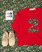 A styled flat-lay of a red long-sleeve children's shirt with a large floral-patterned Liberty Elves number 2 on the front, accompanied by beige lace-up shoes and matching floral hair clips on a green floral background.