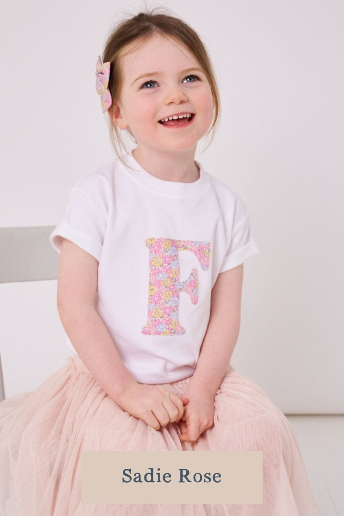 A 5 year old girl wearing a white personalised cotton t-shirt with her initial on the front in Sadie Rose Liberty fabric.